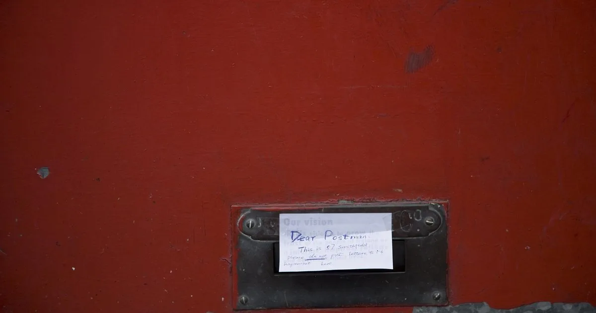 A brass letterbox in a red-painted door, with a handwritten note taped to it asking the postman not to deliver letters addressed to a different building.